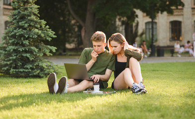 Attentive teen students couple searching content, commenting on laptop sitting on grass in university park. Concentrated students looking at laptop screen, examining information in college garden.