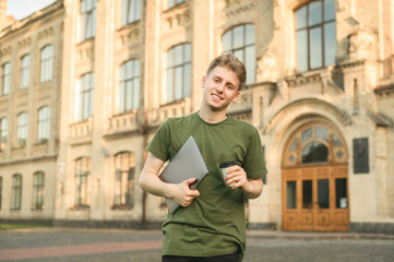 Smiling young student man holding laptop computer looking at camera on a university background. Confident young smiling male student outdoors near college campus with take away coffee and notebook.