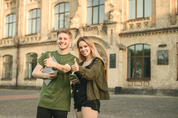 Obraz premium Happy successful students couple smiling showing thumbs up sign at the camera near the college campus. Two cheerful students raising thumbs up near the uni holding laptop, coffee and smartphone.