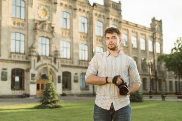 Fototapeta premium Professional male photographer with a beard holding camera standing on the architecture background in the park looking at camera. Handsome young man with camera is satisfied working as a photographer.