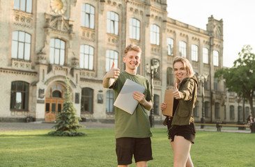 Fototapeta premium Two cheerful students with broad smiles raising thumbs up feeling successful. Exams are over, good grades, cheerful students near the university in the park with laptop and notebook. Like concept.