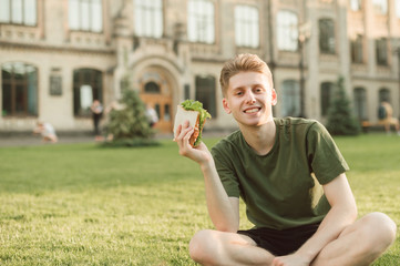 Close photo happy guy in a green T-shirt sits on lawn on the background of the university building with a sandwich in his hands, looking in camera and smiling.Happy student eats a sandwich at a break.