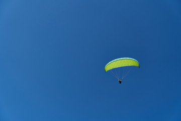 A paraglider with a yellow-green parachute sailing accros a clear, cloudless blue sky. Extreme, adrenaline sports. A lot of negative space for messages.