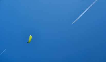 A paraglider with a yellow-green parashute and an airplane leaving a white trail on the blue sky.