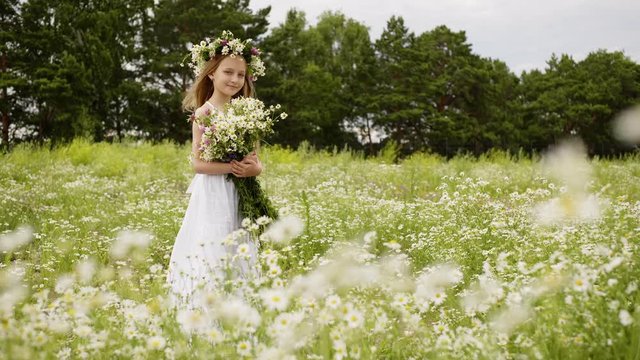 Girl is standing on flower glade looking at camera and smiling. Cute teen girl in white dress with wreath on head and bouquet of daisies at flower meadow. She is posing at shoot. Windy summer day.