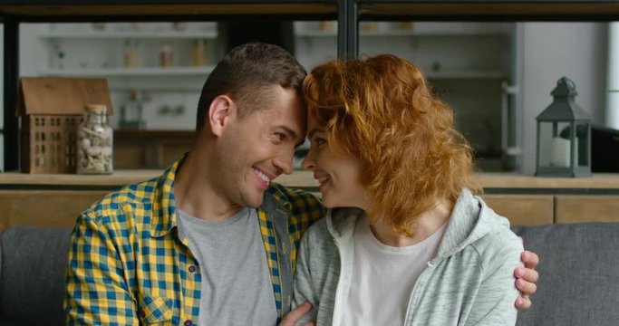 Portrait of young man and woman in love, looking at each other, bowed head to head, smiling, hugging, being happy, enjoying their relationships, sitting on couch at home. 4K, shot on RED camera.