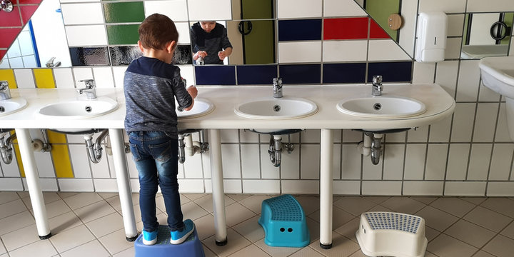 Little caucasian boy washes his hands himself in children's toilet in kindergarten,standing with his back to the camera