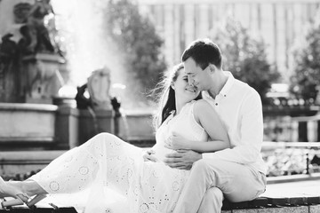 Young couple sitting on a bench at the fountain. A man gently embraces his pregnant wife. Germany, Leipzig