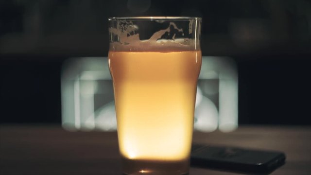 Front view on a glass of craft fresh draft beer stands on the table and glows yellow in backdrop. Shallow depth of field. Blurred background. Slow motion. Camera moves from up to down