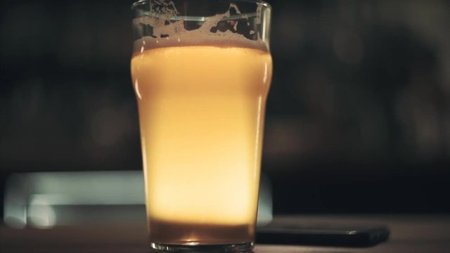 Front view on a glass of craft fresh draft beer stands on the table and glows yellow. Shallow depth of field. Blurred background. Slow motion front view
