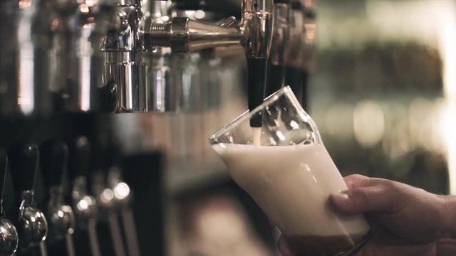 Hand holding a glass and pour nitrogen beer in the craft pub from the tap. Close-up front view. Blurred background in bokeh. Slow motion