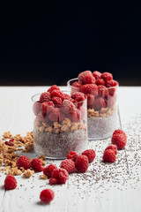 glasses with yogurt, oat flakes and raspberries near scattered chia seeds, oat flakes and berries on white wooden table isolated on black