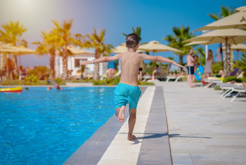 Smiling Caucasian boy having fun in swimming pool at resort on family vacation. He is running along...