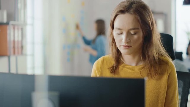 Beautiful and Smart Red Haired Female Specialist Sitting at Her Desk Works on a Desktop Computer. Bright and Modern Open Space Office with Stylish Ergonomic Furniture for Talented Creative People