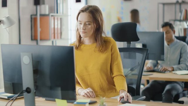Beautiful and Smart Red Haired Female Specialist Sitting at Her Desk Works on a Desktop Computer. Bright and Modern Open Space Office with Stylish Ergonomic Furniture for Talented Creative People