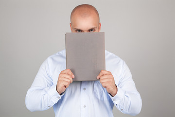 Young guy holding a book in front of his face while wearing a blue shirt, standing on white studio background
