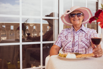 Happy senior woman grandmother outdoor in the terrace sitting at table. Red wineglass in the hand and enjoying some appetizer. Smiles and relax under a pink summer hat