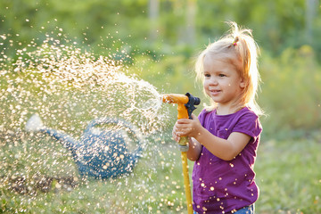 Cute little girl watering flowers in the garden at summer day. Child using garden hose on sunny...