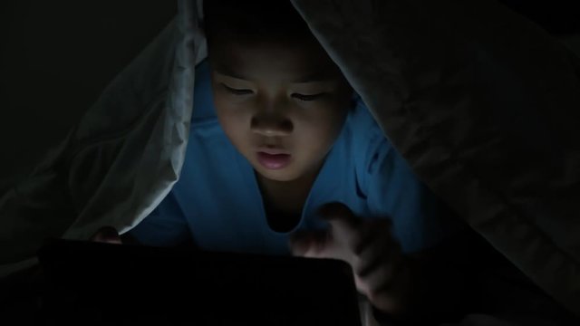 Little boy using tablet under blanket at night in bed.