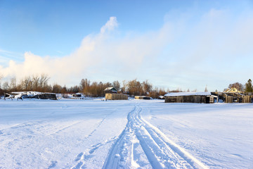 Fototapeta na wymiar Road on snow and ice of frozen lake, tracks going to village, sunny cold weather