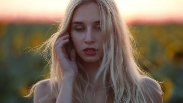 Portrait of young pretty woman in the field at sunset looking at camera, wind blows her blonde hair