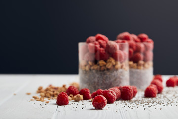 selective focus of scattered raspberries, chia seeds and oat flakes near glasses with yogurt and berries on white wooden table isolated on black
