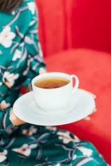 Female hands holding white cup of tea on a red sofa background. Copy space