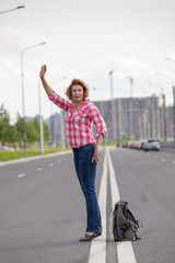 Middle age woman hail a taxi on road, stands on lane with backpack on asphalt