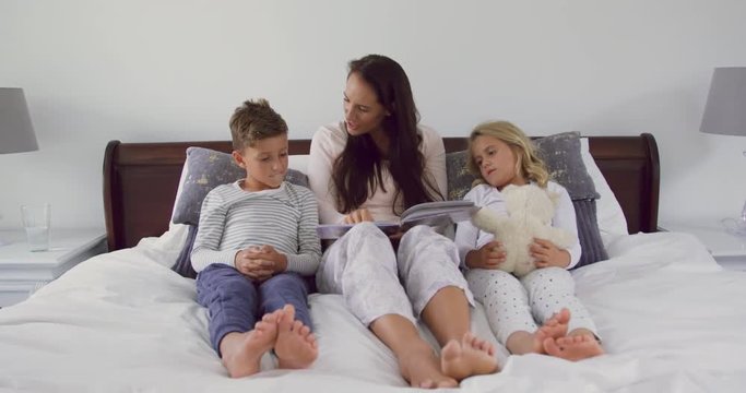 Mother with her children reading story book in bedroom at home 4k