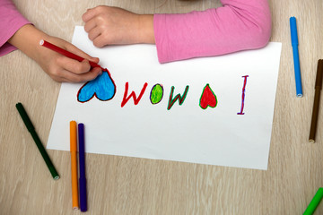 View from above of childs hands drawing with colorful crayons I love Mom on white paper. Art education, creativity concept.