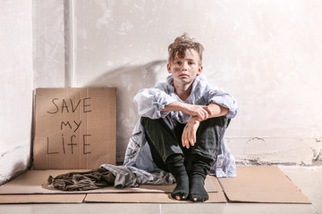 Homeless little boy sitting on floor near wall