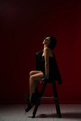 Sensual woman brunette in black tight dress, leather jacket and big brutal boots enjoying resting on a stepladder on red