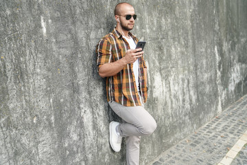 young good looking bold bearded guy standing outdoors against grey modern loft wall with cell phone in his hand chatting and listens music in headphones. wearing yellow shirt, jeans and sunglasses