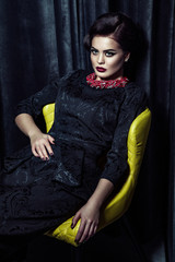 gorgeous brunette woman with stylish makeup and hairdo wearing black evening dress posing while sitting on yellow chair . indoor, studio shot.