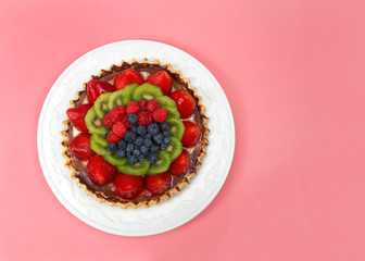 Fresh summer fruit tart with strawberries, kiwi, blueberries, raspberries on a white porcelain plate with pink background. Top view flat lay.