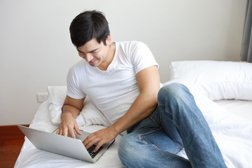 Fototapeta premium portrait of handsome caucasian in white shirt and blue jean using computer on bed in bedroom with relaxation and smiling face