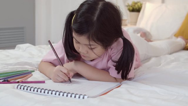 Young Asian girl drawing at home. Asia japanese woman child kid relax rest fun happy draw cartoon in sketchbook before sleep lying on bed, feel comfort and calm in bedroom at night concept.