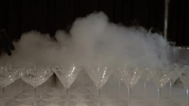 Cold, white smoke spread along a row on the table, empty Martini glass. Leaving the camera from a large number of glasses shrouded in white smoke