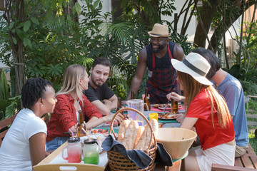 Group of happy friends eating and drinking beers at barbecue dinner camping in nature and having meal together outdoor as summer lifestyle, food and friendship concept