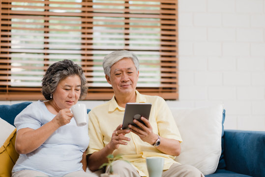 Asian elderly couple using tablet and drinking coffee in living room at home, couple enjoy love moment while lying on sofa when relaxed at home. Enjoying time lifestyle senior family at home concept.
