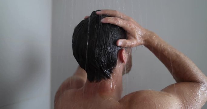 Man in shower washing hair showering in bathroom at home. Unrecognizable person from behind rinsing shampoo and conditioner from hair in warm bath with modern bathroom.