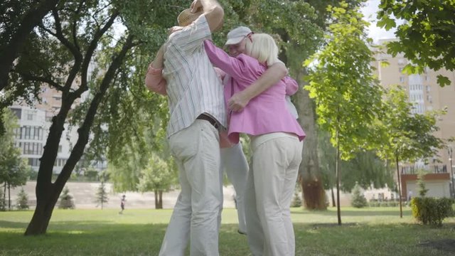 Two cute adult couples hugging in the park together standing in circle. Double date of senior couples. Friendly company resting outdoors. Old men and women smiling happily. Slow motion.