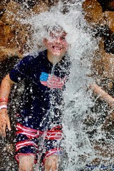 young boy stands in a waterfall at a pool.