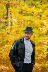 Smiling man in black hat, leather jacket, white shirt and dark trousers on yellow autumn...