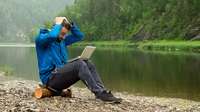 A guy in a blue jacket with a white laptop is sitting on a log in the rain on the bank of the river and holding his head. Conceptual error in programming or disruption of a business transaction.
