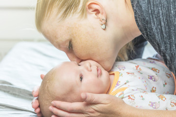 Woman is stroking and kissing newborn baby lying on blanket. Baby is looking at the side.