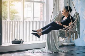 An elderly woman with a Cup in her hands listening to a retro player, sitting in a swing chair by...