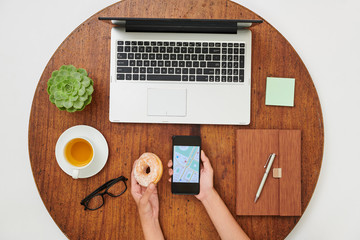 Naklejka premium Close-up of woman sitting at wooden table in front of laptop computer drinking tea and eating doughnut while watching online city map on mobile phone