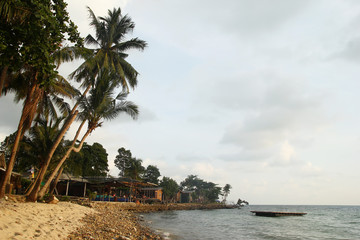 View on a sandy and stony beach with high palm trees and wooden houses near to the sea at sunset. Koh Chang, Thailand.