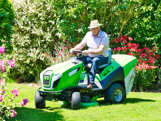 Senior man driving a tractor lawn mower in garden with flowers